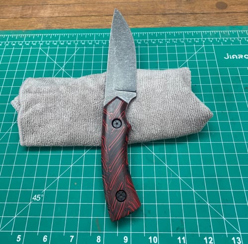 Customer's knife with a textured red and black handle, resting on a gray cloth atop a green cutting mat.