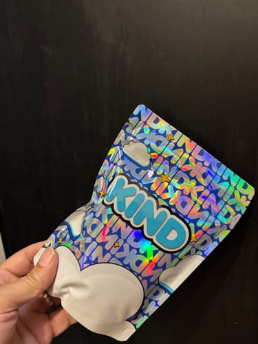 Customer's hand holding a holographic blue KIND White Widow pouch against a dark background.