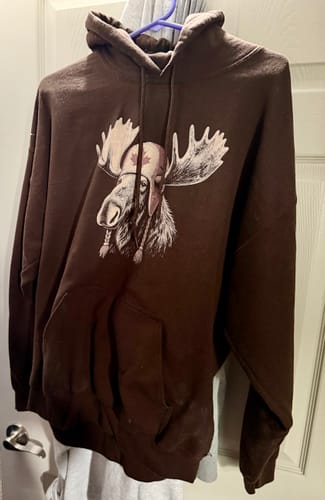 Customer's brown Canada Moose Cap Hoodie hanging on a hanger, featuring a moose graphic in a Canadian cap.