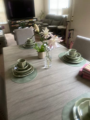 Customer's Royal Olive 16-Piece Organic Dinnerware Set arranged on a wooden dining table with a vase of flowers.