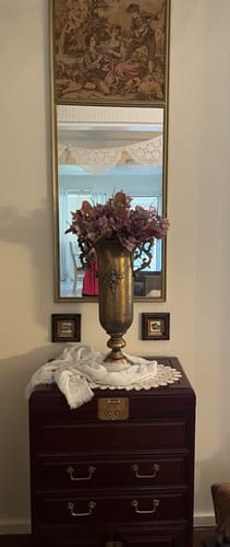 Customer's Coralie French Provincial Urn Vase in aged gold metal, filled with flowers on a dark wood cabinet.