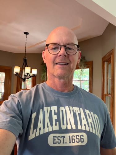 Customer's Lake Ontario Varsity T-Shirt in blue on a smiling person indoors.