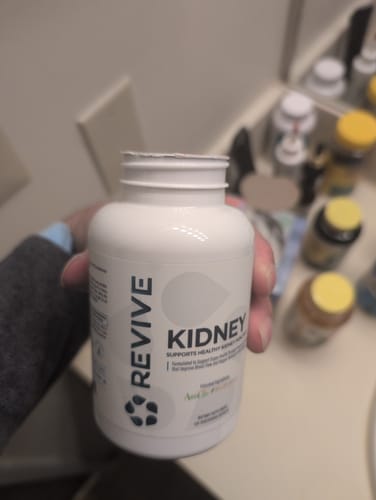 Customer's hand holding a white bottle of Revive Kidney supplement in a bathroom setting.