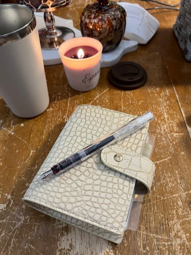 Customer's closed ivory crocodile-embossed Ivory Safari planner with a clear pen on a rustic wooden table.