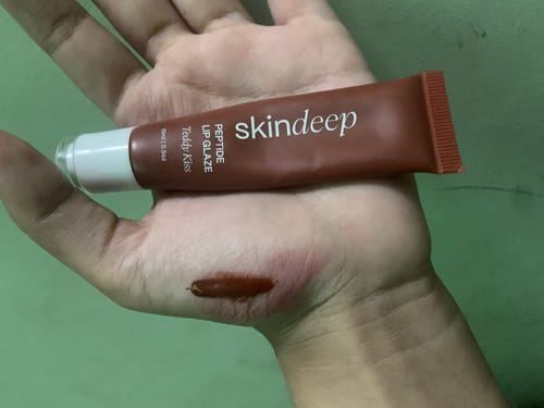 Customer's hand holding a tube of Peptide Lip Glaze with a brown swatch of the product on their palm.
