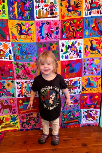 Customer's toddler wearing the black Toddler Chingona Como Mi Abuela T-Shirt, standing before a colorful textile.