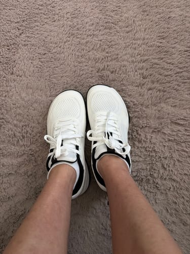 Customer's feet wearing white CUURT Muuv wide toe box court shoes while standing on a textured beige rug.