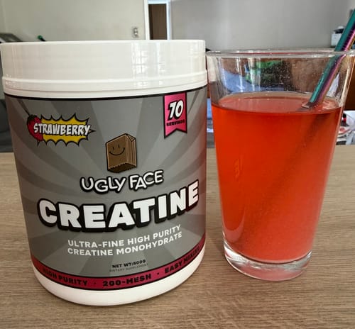 Customer's UglyFace Creatine tub next to a glass of mixed red liquid with a reusable straw on a wooden table.