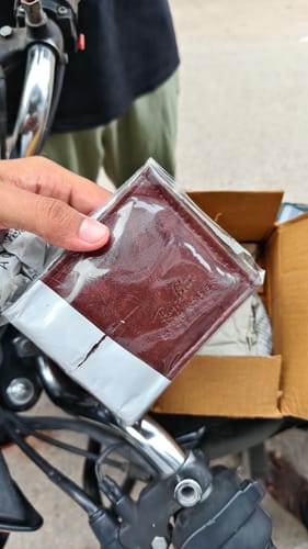 Customer's hand holding a brown wallet in plastic packaging over a cardboard box.