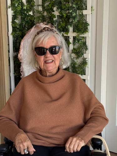 Customer's Zen Rib Dolman Sweater in deep camel, worn by a reviewer in a wheelchair against a green vine backdrop.