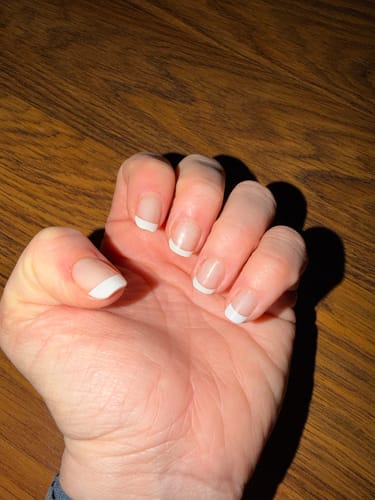 Hand einer Kundin, die die aufgetragenen 'Classy French' UV-Gel-Nagelfolien mit klassischen weißen Spitzen auf kurzen Nägeln zeigt.