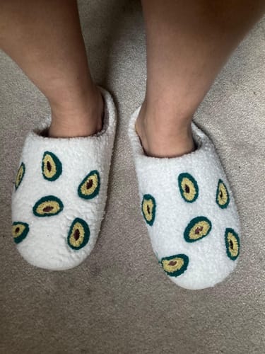 Customer's feet in plush, white Avocado Plush Slippers featuring a repeating avocado pattern.