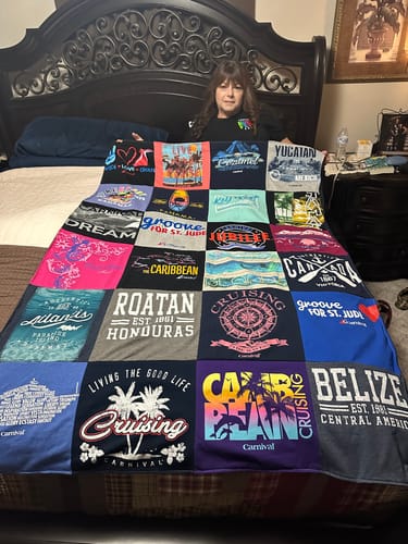 Customer holding up their finished Premium T Shirt Quilt, made of many colorful shirts, while sitting on a bed.