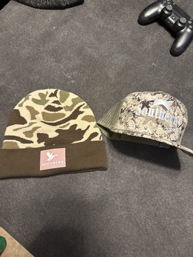 Customer's camouflage beanie and matching baseball cap from the Hat of the Month subscription laid on a dark surface.