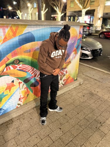 Customer wearing a brown Goat Hoodie with a white logo, posing in front of a colorful mural at night.