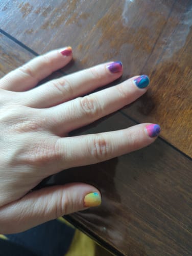 Hand einer Kundin, die die bunten „Rainbow Smoke“-Nagelfolien auf kurzen Nägeln trägt.