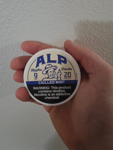 Reviewer holding a round, white tin of ALP Chilled Mint Nicotine Pouches against a plain wall.