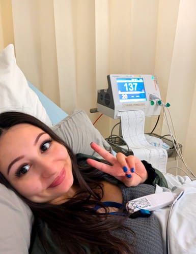 Reviewer in a hospital bed wearing full Double Trouble Length lashes, smiling and giving a peace sign.
