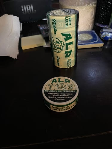 Customer's can and 5-pack sleeve of Mountain Wintergreen Nicotine Pouches displayed on a dark desk.