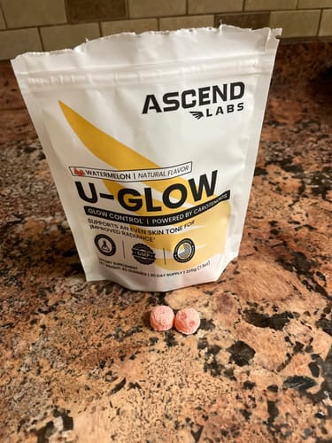 Reviewer's U-GLOW supplement pouch and two pink chewable tablets on a counter.