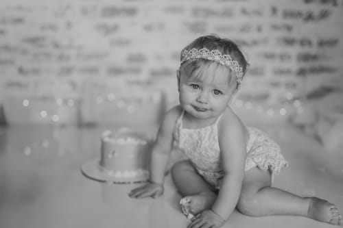 Customer's baby wearing a lacy outfit and matching headband while sitting next to a small cake.