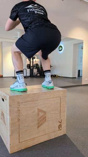 Kunde in Hockposition auf der hölzernen Plyobox in einem Fitnessstudio.