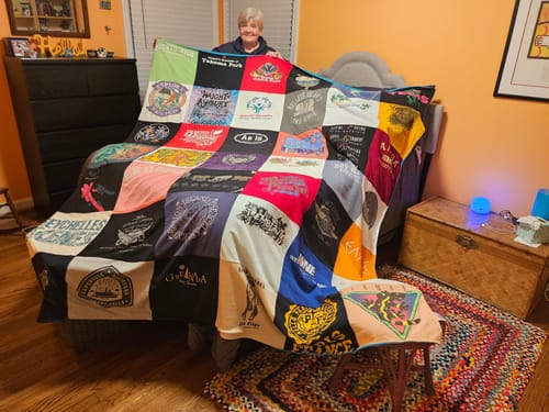 Customer holding up a large Premium T Shirt Quilt made of colorful, unique t-shirt squares in a bedroom.