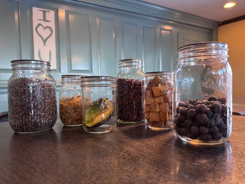 Customer's Chalkboard Top Mason Jar Lids on six glass jars filled with various foods, including fresh blueberries, on a counter.
