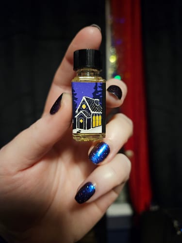 Reviewer's hand with dark sparkly nails holding a small bottle of A Confection Of Sorrows, showing its illustrated gingerbread house label.