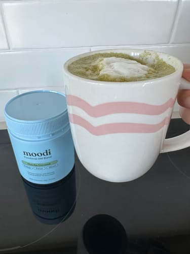 Reviewer holding a mug of their prepared, frothy Matcha Coconut drink, with the product tub on the counter.