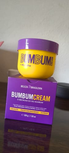 Cliente exibe o Bumbum Cream, um pote amarelo com tampa roxa, sobre a sua caixa roxa.