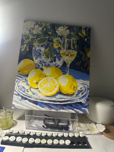 Reviewer's completed 'Lemons and Wine' paint by numbers artwork on a desk with the kit's paint pots shown in front.