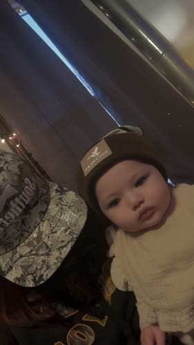 Reviewer and a baby wearing two hats from the Hat of the Month Crew, one a camouflage baseball cap and the other a dark beanie.