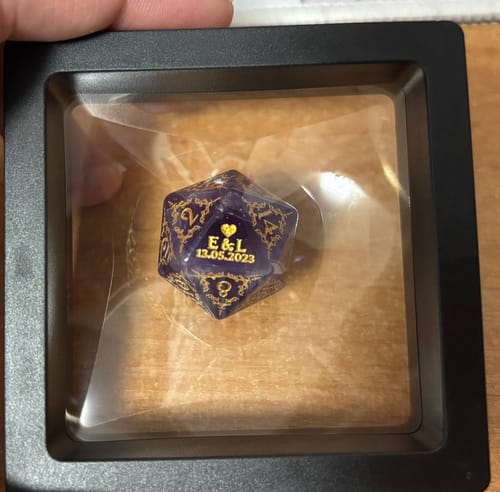 A customer's custom purple D20 die in its black display box, showing personalized gold engraving with initials and a date.
