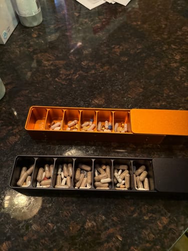 Customer's orange and black Weekly AM-PM Vitamin XL Cases open on a dark counter, showing the compartments filled with pills.