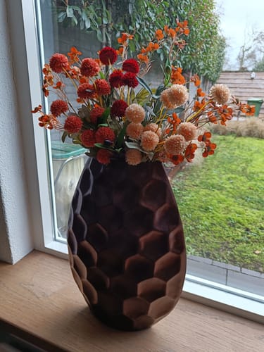 Recensent toont het Wax Arrangement met oranje en rode tinten in een donkere, getextureerde vaas op de vensterbank.