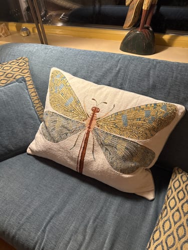 Recensent toont het grijsblauwe jacquard kussen met libel op een blauwe bank.