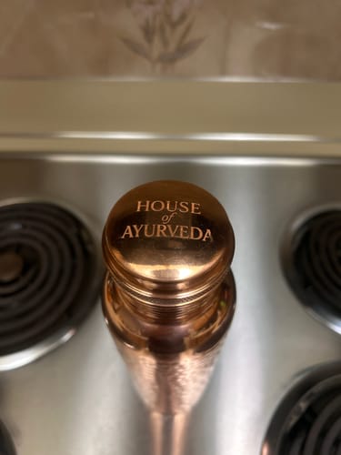 Close-up: Reviewer's copper water bottle showing the cap engraved with 'House of Ayurveda' on a kitchen stovetop.