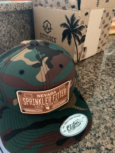 Customer's camouflage hat with the Nevada Engraved License Plate leather patch on the front, sitting on a counter.