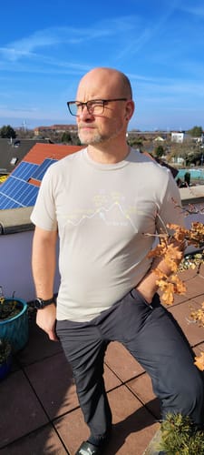 Kunde trägt das Bio Herren Shirt '5 Phasen der Wanderung' bei sonnigem Wetter auf einer Terrasse.