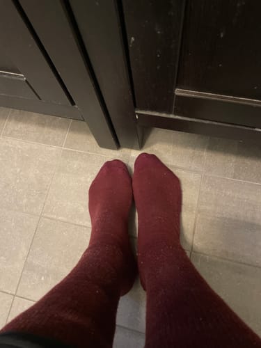 Recensent som visar sina bekväma, mörkröda Diasocks Komfortstrumpor på fötterna inomhus.