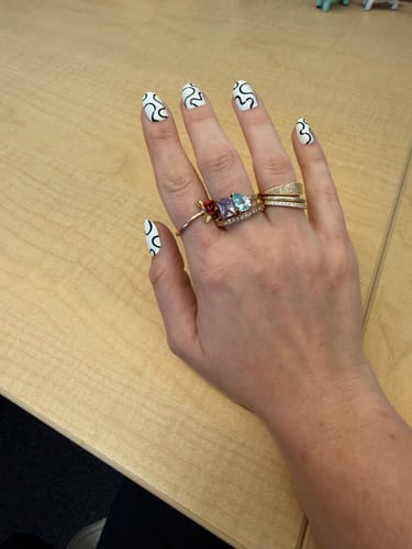Reviewer's hand showing the Toi Et Moi Birthstone Ring with multiple colorful stones, stacked with other rings on a wooden desk.