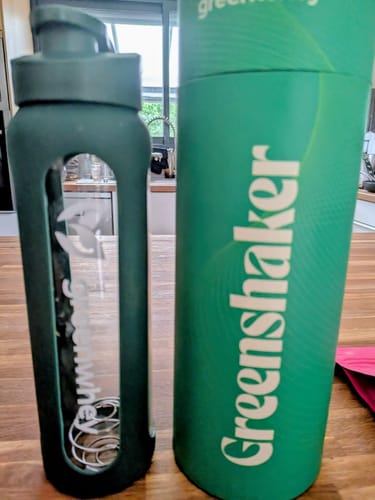 Avis d'un client: le GreenShaker 2.0 vert à côté de son bel emballage cylindrique sur une table en bois.