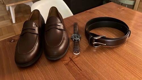 Cliente muestra su Cinturón Formal Brandy sobre una mesa de madera, a juego con zapatos y un reloj de cuero.