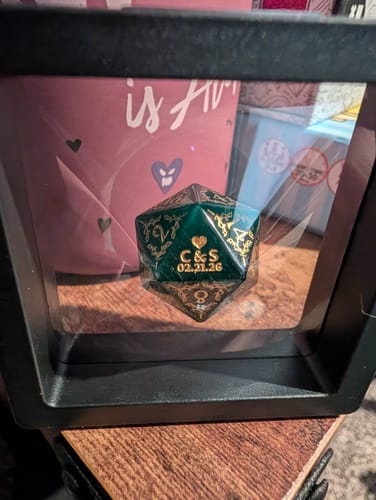 Customer's personalized green D20 dice in a black display frame. The '20' face shows the custom engraving 'C&S 02.21.26'.