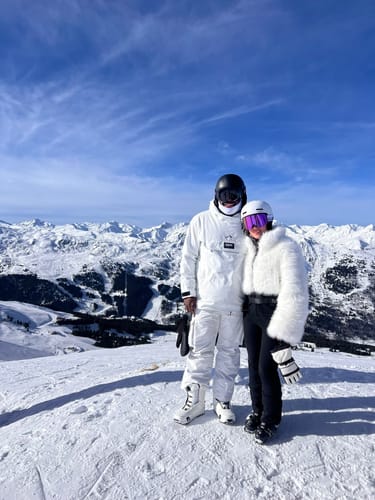 Customer on a snowy mountain wearing the ski suit's white faux fur jacket and black pants.