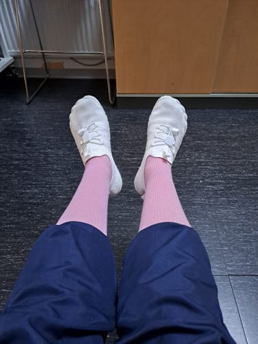 Arvostelijan jalassa valkoiset Vapauta Varpaat -paljasjalkakengät vaaleanpunaisten sukkien kanssa sisätiloissa.