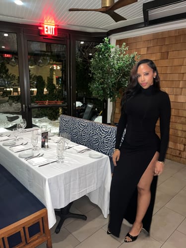 Reviewer in a restaurant wearing the black turtleneck Butter Contour Maxi Dress with a high thigh slit.