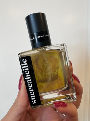 Reviewer holding a square bottle of Liquid Gold perfume, showing the shimmery golden liquid inside.