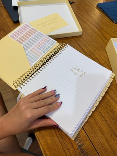 Reviewer's hand on the open Butter Yellow 2026 Daily Planner, showing the gold spiral binding and included sticker sheets.